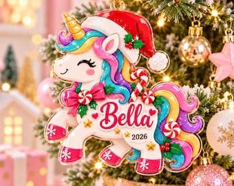 Op de afbeelding: Een platte kerstboomversiering van acryl in de vorm van een eenhoorn met een rode kerstmuts. De eenhoorn heeft manen in regenboogkleuren, een gouden hoorn en roze hoeven met witte sneeuwvlokken. De naam Bella en 2026 staan in rode letters.