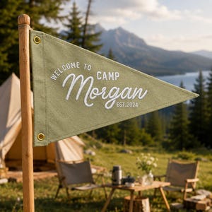 May include: A triangular olive green canvas pennant flag attached to a wooden pole. The flag features white embroidered text that reads Welcome to Camp Morgan Est. 2024. It includes two gold-colored metal grommets along the left vertical edge.