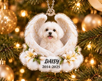 May include: A clear acrylic Christmas ornament featuring a white dog framed by two large angel wings. Below the dog are white lily flowers and the text Daisy 2014-2025. The ornament hangs from a silver beaded chain against a blurred tree.