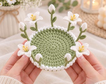 May include: A round, sage green crocheted coaster featuring a white scalloped border. The coaster is decorated with several small, three-dimensional white lily flowers and closed lily buds on green stems attached to the outer edge.
