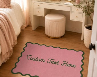 May include: A rectangular pink rug with a scalloped dark green border. The rug features the text Custom Text Here in a dark green cursive font centered on the surface. The rug is positioned on a light brown wooden floor in a bedroom setting.