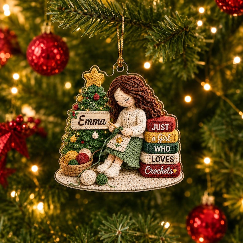 May include: A clear acrylic Christmas ornament featuring a crocheted figure with brown hair sitting next to a green Christmas tree and a stack of books. The tree has a sign reading Emma. The books read JUST a Girl WHO LOVES Crochets.