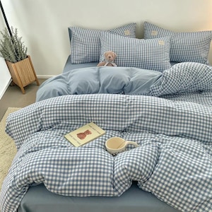 Peut inclure: Un ensemble de lit composé d'une housse de couette et de taies d'oreiller à motif vichy bleu clair et blanc. Un petit ours en peluche est posé sur le lit, accompagné d'un livre illustré d'un ours et d'une tasse en céramique blanche.
