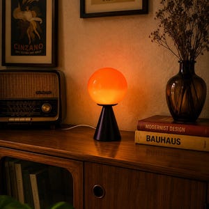 May include: A table lamp featuring a spherical, glowing orange glass shade mounted on a solid black conical base. The lamp emits a warm, diffused light, creating a soft illumination that highlights the smooth, rounded form of the glass globe.