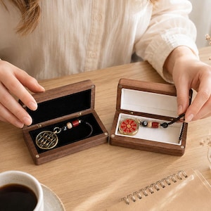 Puede incluir: Dos cajas rectangulares de madera con tapas abatibles, cada una contiene un colgante circular en un cordón de cuentas. Un colgante presenta un diseño de ábaco dorado, mientras que el otro es una pieza circular roja con caracteres chinos dorados.