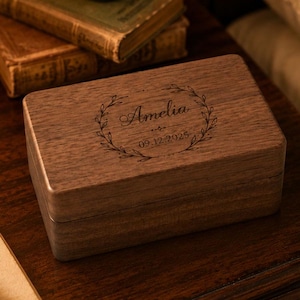 May include: A rectangular dark brown wooden box with a smooth finish. The lid features a laser-engraved design of a circular wreath surrounding the name Amelia and the date 09.12.2025 in a script and serif font combination.