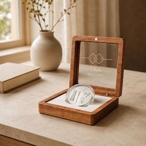 May include: A square wooden display box with a hinged glass lid featuring the engraved text Ethan and March 3, 2025, along with a geometric design. Inside, a circular silver coin with etched details rests in a white insert.