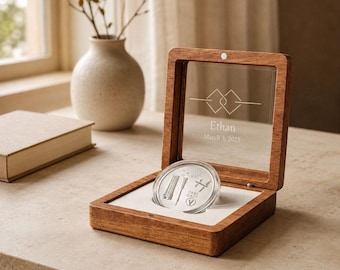 May include: A square wooden display box with a hinged glass lid featuring the engraved text Ethan and March 3, 2025, along with a geometric design. Inside, a circular silver coin with etched details rests in a white insert.
