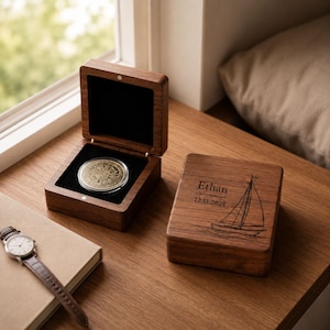 May include: A dark wood square box with a hinged lid, featuring an engraved sailboat, the name Ethan, and the date 12.11.2024. An open matching box contains a circular gold-coloured coin with intricate embossed details set against a black interior.