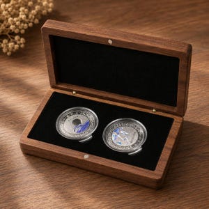 Könnte beinhalten: Eine rechteckige Aufbewahrungsbox aus dunkelbraunem Holz mit Klappdeckel und schwarzem Samtfutter. Im Inneren befinden sich zwei kreisförmige, silberfarbene Münzen, die jeweils in durchsichtigen Schutzkapseln mit blauen Details liegen.