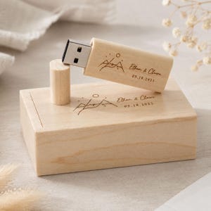 Puede incluir: Una memoria USB de madera clara y una caja rectangular de madera a juego. Ambos artículos presentan un grabado láser de una montaña, los nombres Ethan & Claire y la fecha 05.18.2025. La memoria USB tiene un conector de metal plateado.