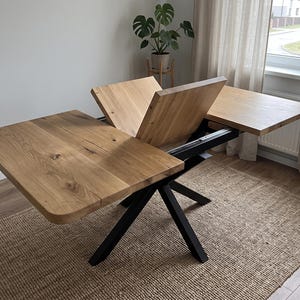 Könnte beinhalten: Ein ausziehbarer Esstisch mit einer Tischplatte aus hellem Naturholz mit sichtbaren Astlöchern und einem schwarzen, X-förmigen Metallgestell. Der Tisch ist teilweise ausgeklappt und zeigt einen klappbaren Mittelteil, der die beiden Hauptplatten verbindet.