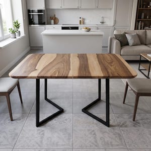 May include: A rectangular dining table featuring a natural wood grain tabletop with light and dark brown tones, supported by two black metal trapezoidal legs. The table is positioned between two light-coloured chairs on a tiled floor.
