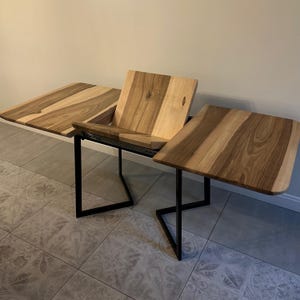 May include: A wooden desk with a natural grain finish featuring two side extensions and a central angled panel. The desk is supported by a black metal frame with a geometric design, providing a stable base for the light and dark brown wood surfaces.