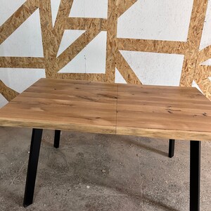 May include: A rectangular dining table featuring a natural light brown wood grain tabletop with a live edge finish. The table is supported by four sturdy, angled black metal legs that provide a stable base for the wooden surface.