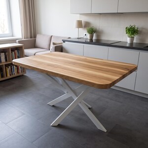 Pode incluir: Uma mesa de jantar retangular com um tampo de madeira castanho-claro natural com veios visíveis e cantos arredondados. A mesa apresenta uma base de metal branco com um design de pernas em forma de cruz que suporta o tampo.