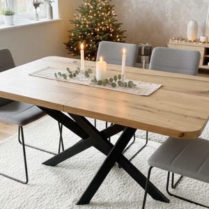 Könnte beinhalten: Ein rechteckiger Esstisch aus hellem Holz mit abgerundeten Ecken und einem schwarzen X-förmigen Metallgestell. Der Tisch ist von vier grauen Polsterstühlen mit dünnen schwarzen Metallbeinen umgeben und hat einen beigen Tischläufer.