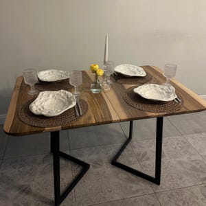 May include: A rectangular wooden dining table with a natural grain pattern and black metal legs. The table is set with four woven brown placemats, four irregular white textured plates, four clear textured glasses, a glass vase with yellow flowers, and a tall white candle.