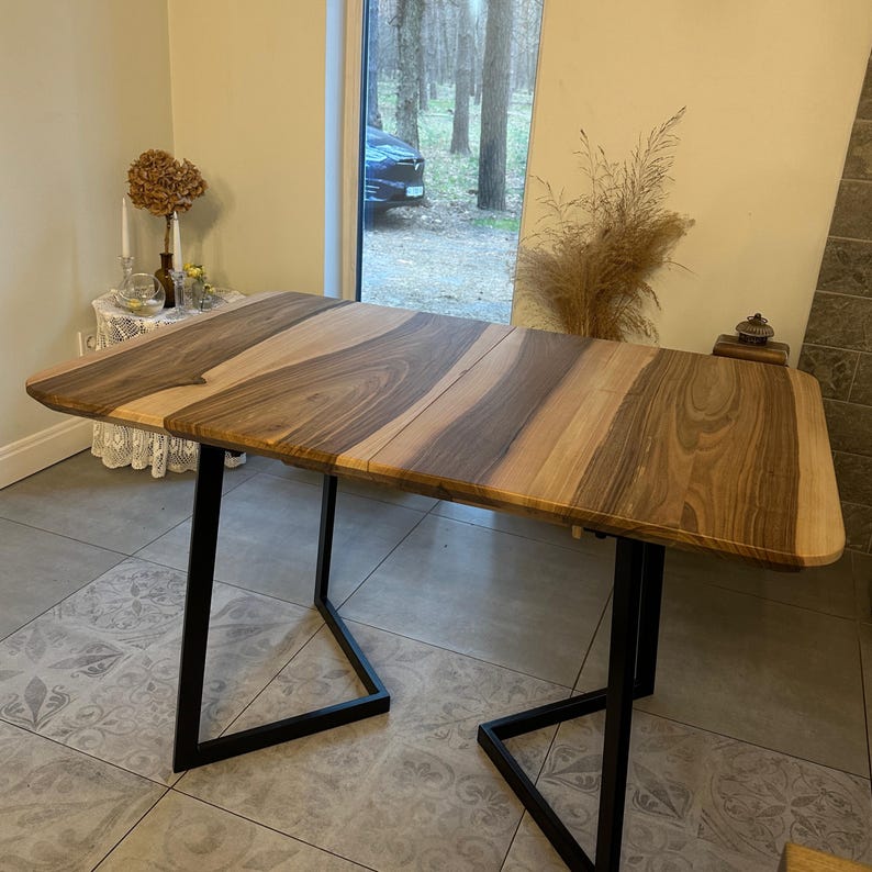 May include: A rectangular dining table featuring a natural wood grain surface with alternating light and dark brown tones. The tabletop is supported by two black metal U-shaped legs, providing a stable base for the smooth, polished wooden surface.