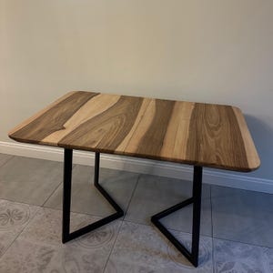 May include: A rectangular table featuring a natural wood grain tabletop with light and dark brown tones. The table is supported by two black metal legs, each constructed in a geometric, open-frame design that provides a stable base for the surface.