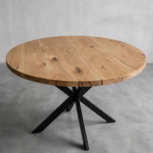 Pode incluir: Uma mesa de jantar redonda com um tampo de madeira castanho-claro natural com nós visíveis e uma costura central distinta. A mesa é suportada por uma base de metal preto composta por quatro pernas cruzadas que formam um formato de X.