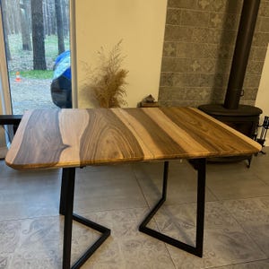 May include: A rectangular table featuring a natural wood grain top with contrasting light and dark brown tones, supported by a black metal frame with two U-shaped legs. The surface displays distinct wood patterns and smooth, rounded corners.