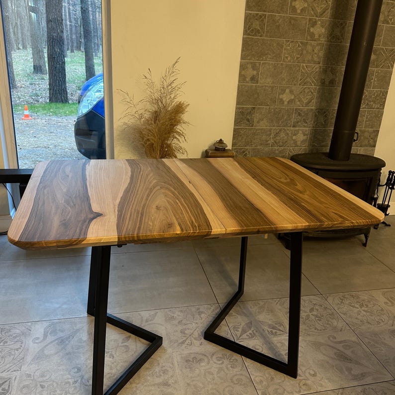 May include: A rectangular table featuring a natural wood grain top with contrasting light and dark brown tones, supported by a black metal frame with two U-shaped legs. The surface displays distinct wood patterns and smooth, rounded corners.