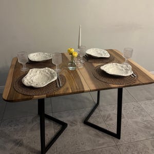 May include: A rectangular dining table with a natural wood grain top and black metal legs. The table is set for four with textured brown placemats, irregular white ceramic plates, clear glass goblets, silver cutlery, and a central glass vase with yellow flowers.
