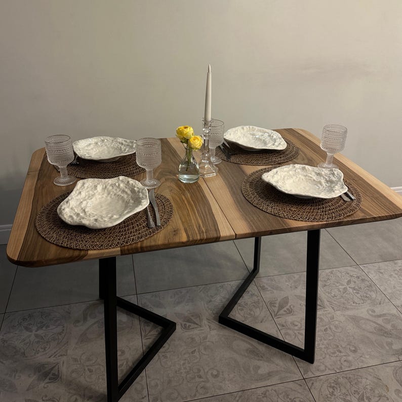 May include: A rectangular dining table with a natural wood grain top and black metal legs. The table is set for four with textured brown placemats, irregular white ceramic plates, clear glass goblets, silver cutlery, and a central glass vase with yellow flowers.