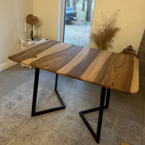 May include: A rectangular dining table featuring a natural wood grain surface with alternating light and dark brown tones. The tabletop has rounded corners and is supported by two black metal U-shaped legs positioned at each end of the table.