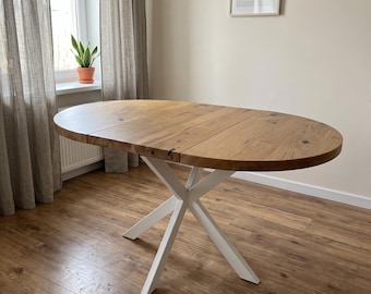 Könnte beinhalten: Ein ovaler Esstisch mit einer Tischplatte aus naturfarbenem, hellbraunem Holz mit sichtbaren Maserungen und Astlöchern. Der Tisch steht auf einem weißen Metallgestell, das aus vier schrägen Beinen besteht, die sich unter der Platte treffen.