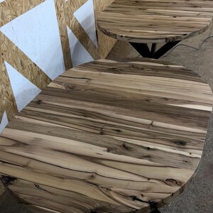 K&ouml;nnte beinhalten: Zwei runde Esstische mit nat&uuml;rlicher Holzmaserung in dunkelbraunen und hellbeigen Streifen. Jeder Tisch hat eine glatte, kreisf&ouml;rmige Oberfl&auml;che und wird von einer schwarzen Metallunterkonstruktion getragen, die unter den Kanten sichtbar ist.