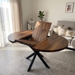 May include: A round wooden dining table with a dark brown walnut grain finish and a central leaf extension. The table features a black metal base with intersecting legs and is shown in a partially extended position with the leaf angled upwards.