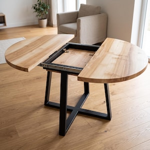 Pode incluir: Uma mesa de jantar redonda de madeira com acabamento em grão de madeira natural castanho claro e uma base de metal preta. A mesa apresenta um design extensível com duas secções superiores semicirculares que se separam.