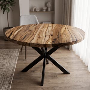 Puede incluir: Una mesa de comedor redonda con un tablero de madera natural que presenta vetas y nudos oscuros prominentes. La mesa se apoya sobre una base de metal negro compuesta por cuatro patas entrecruzadas que se unen en el centro de la estructura.