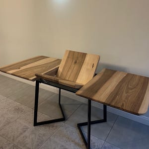 May include: A convertible table featuring a natural wood grain surface with light and dark brown tones. The table has a central section that can be raised or lowered, supported by a black metal frame with angular, geometric legs on each side.