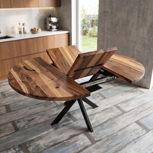 Puede incluir: Una mesa de comedor redonda de madera con acabado de veta natural que cuenta con un mecanismo de extensión central. La mesa tiene una base de metal negro con patas anguladas y se muestra parcialmente abierta, revelando los rieles deslizantes.