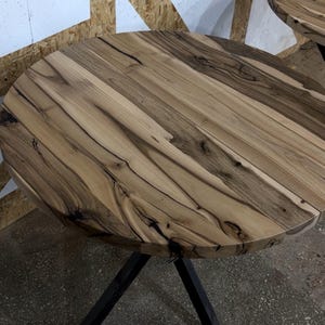 K&ouml;nnte beinhalten: Ein runder Tisch mit einer Tischplatte aus Naturholz, die helle beige und dunkelbraune Maserungen, Astl&ouml;cher und organische Muster aufweist. Die Tischplatte wird von einem schwarzen Metallgestell mit schr&auml;gen Beinen getragen.