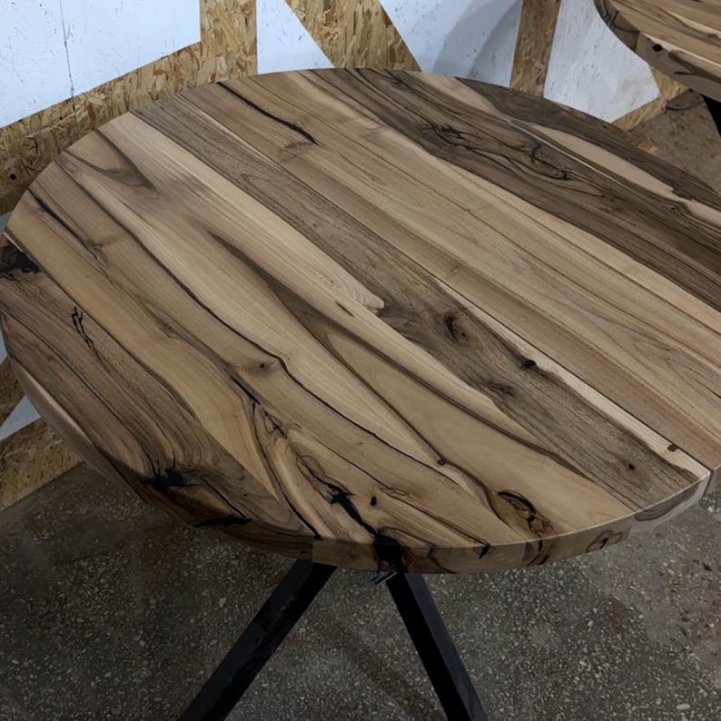 K&ouml;nnte beinhalten: Ein runder Tisch mit einer Tischplatte aus Naturholz, die helle beige und dunkelbraune Maserungen, Astl&ouml;cher und organische Muster aufweist. Die Tischplatte wird von einem schwarzen Metallgestell mit schr&auml;gen Beinen getragen.