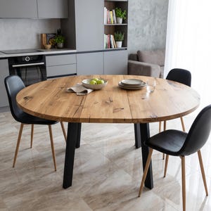 Pode incluir: Uma mesa de jantar redonda com tampo de madeira castanho-claro natural com veios visíveis e quatro pernas de metal preto, acompanhada por quatro cadeiras pretas com pernas de madeira clara. A superfície da mesa apresenta nós visíveis.