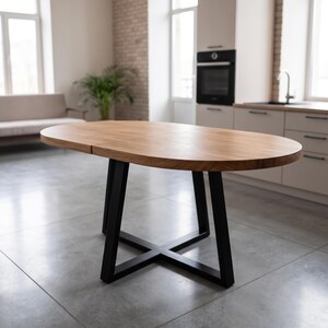 Puede incluir: Una mesa de comedor ovalada con una superficie de madera de color marrón claro y un patrón de veta visible. La mesa se apoya sobre una base de metal negro construida con cuatro patas anguladas que se unen en forma de X en el suelo.