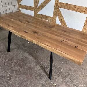May include: A rectangular dining table featuring a natural light brown wood grain surface with visible knots and a live edge. The table is supported by four straight, matte black metal legs positioned at each corner of the rectangular frame.