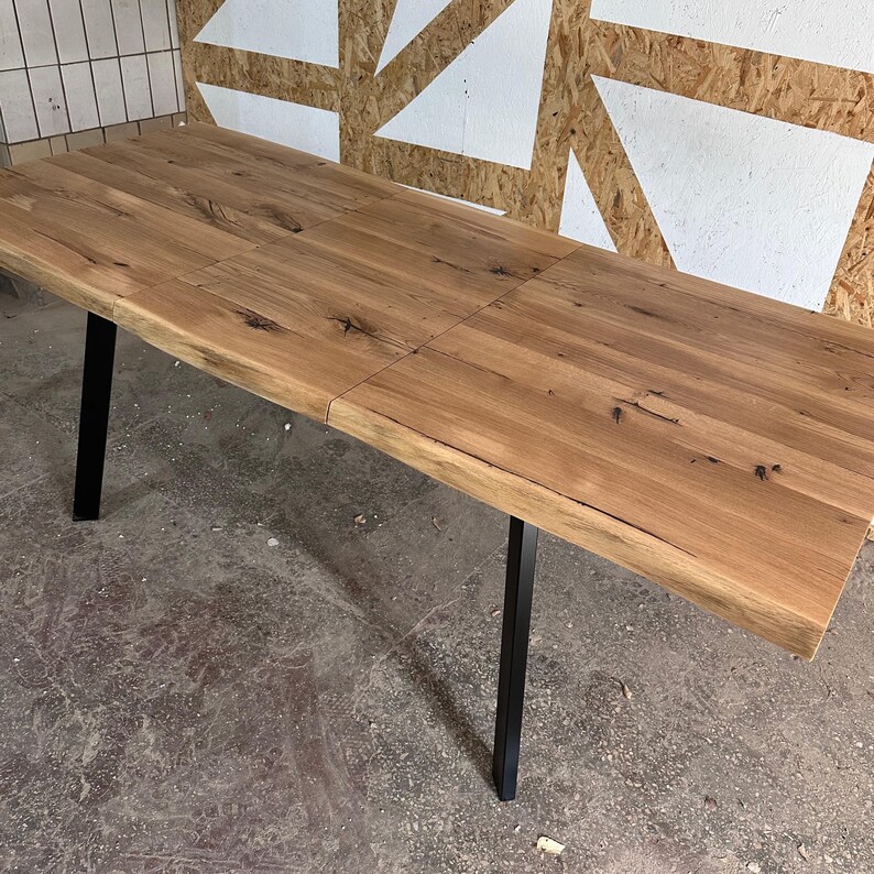 May include: A rectangular dining table featuring a natural light brown wood grain surface with visible knots and a live edge. The table is supported by four straight, matte black metal legs positioned at each corner of the rectangular frame.