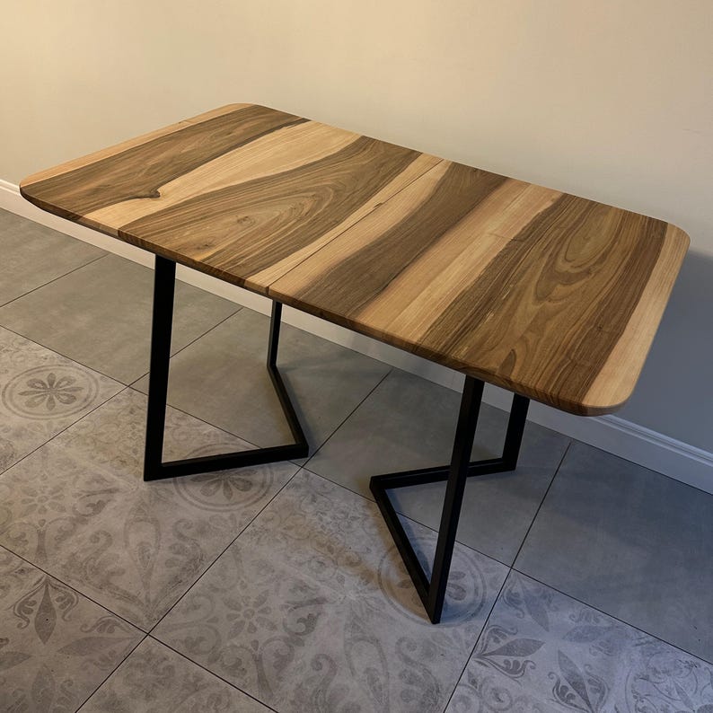 May include: A rectangular table featuring a natural wood grain top with light and dark brown tones and rounded corners. The table is supported by two black metal legs, each constructed in a geometric, angular frame design that provides stability.