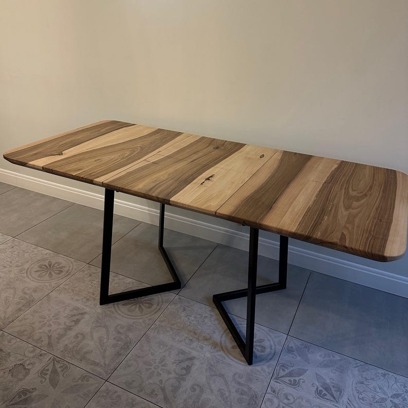 May include: A rectangular dining table featuring a natural wood grain tabletop with alternating light and dark brown tones. The table is supported by two black metal legs, each designed with a geometric, angular frame that provides a stable base.