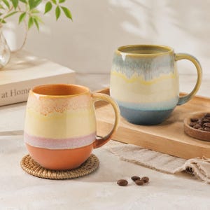 May include: Two ceramic mugs with rounded bodies and handles. The front mug features horizontal bands of orange, pink, yellow, and cream. The back mug displays a gradient of blue, yellow, and green tones. Both mugs have a smooth, glossy finish.