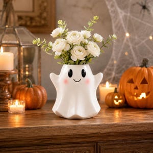 May include: A white ceramic ghost-shaped vase with a smooth finish, featuring two black oval eyes, a small curved mouth, and soft pink circular cheeks, filled with a bouquet of white ranunculus flowers and green foliage stems.