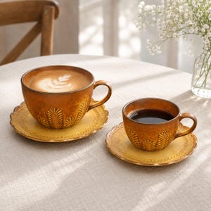 Puede incluir: Dos tazas de cerámica con platillos en un esmalte cálido de color ámbar y amarillo. Las tazas presentan un patrón texturizado y en relieve de hojas tipo helecho en el exterior. Una taza contiene un café con leche con arte en espuma y la otra café negro.