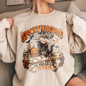 May include: A beige crewneck sweatshirt featuring a graphic print of a black cat wearing a chef hat and apron, baking ghost-shaped cookies. The text reads "SPOOKY BISCUITS BAKERY" and "WE KNEAD EM YOU NEED EM" with illustrations of a pumpkin and flour.