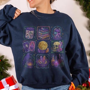 May include: A navy blue long-sleeve sweatshirt featuring a grid of nine colorful illustrations including a jar of eyeballs, a stack of books, mushrooms in a glass dome, tarot cards, a crystal ball, a skull, a raven with a lantern, crystals, and potions.